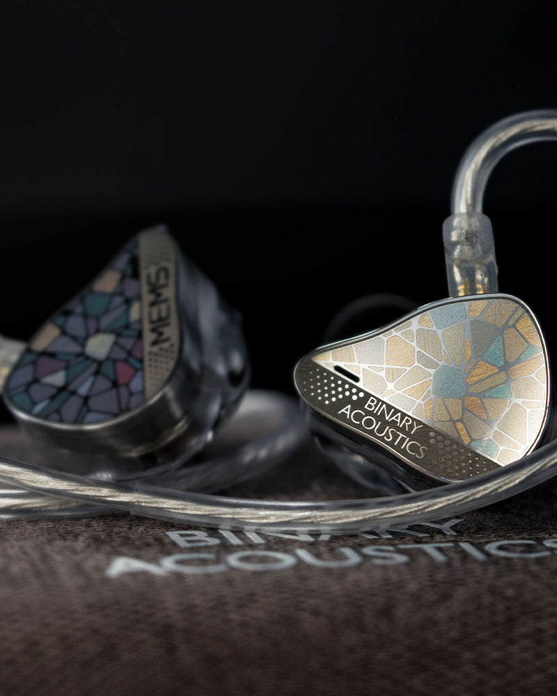 Close-up of Binary Acoustics MEMS in-ear monitors with a mosaic iridescent finish on the ear tips and a clear cable, resting on a dark textured surface with the Binary Acoustics logo visible.