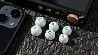 Kiwi Ears Flex Ear Tips, six grey tips on black foil next to a phone and a music player