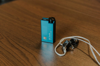 iBasso DC-Tonfa standing on a wooden desk next to a pair of IEMs with a silver cable
